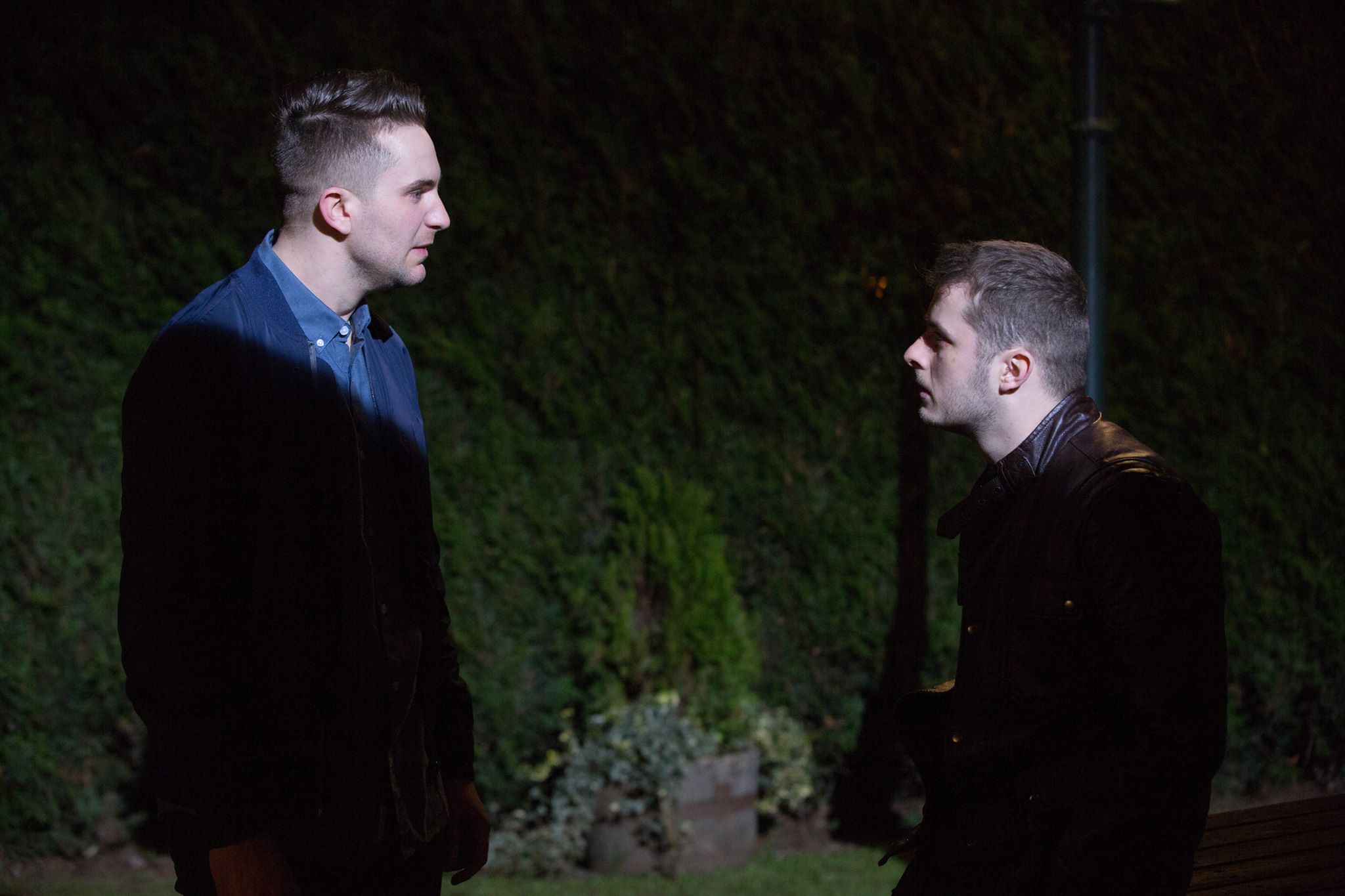Ben and Callum from EastEnders, standing in the park with only their faces illuminated, looking at each other